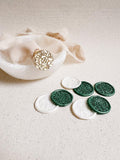 Brass wax stamp with an intricate clover field design placed on top of a small frosted glass bowl with a ribbon on top. In the foreground are wax seals made with the stamp in green and pearl.