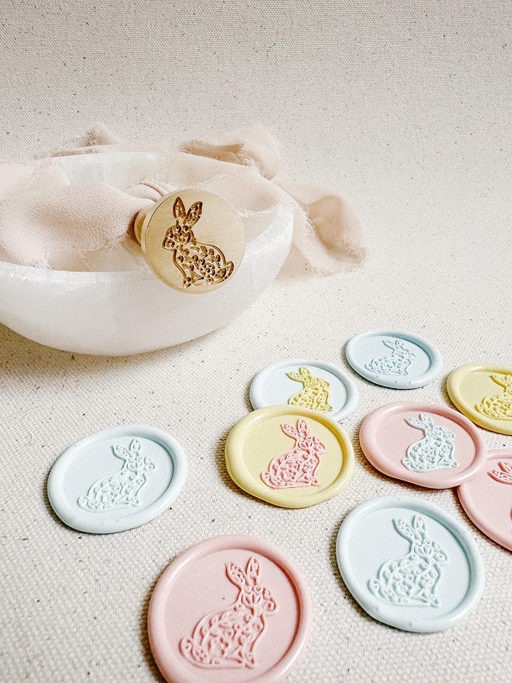 brass wax stamp engraved with small floral designs within the silhouette of a bunny placed on a small frosted glass bowl and a ribbon on top. In the foreground are bunny wax seals made with the stamp in mixed colours, including yellow, pink and blue.