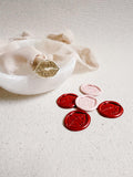 Wax stamp in the shape of lips placed on top of a frosted glass bowl with a delicate ribbon on top. In the foreground are 5 wax seals in the shape of lips in pink and red.